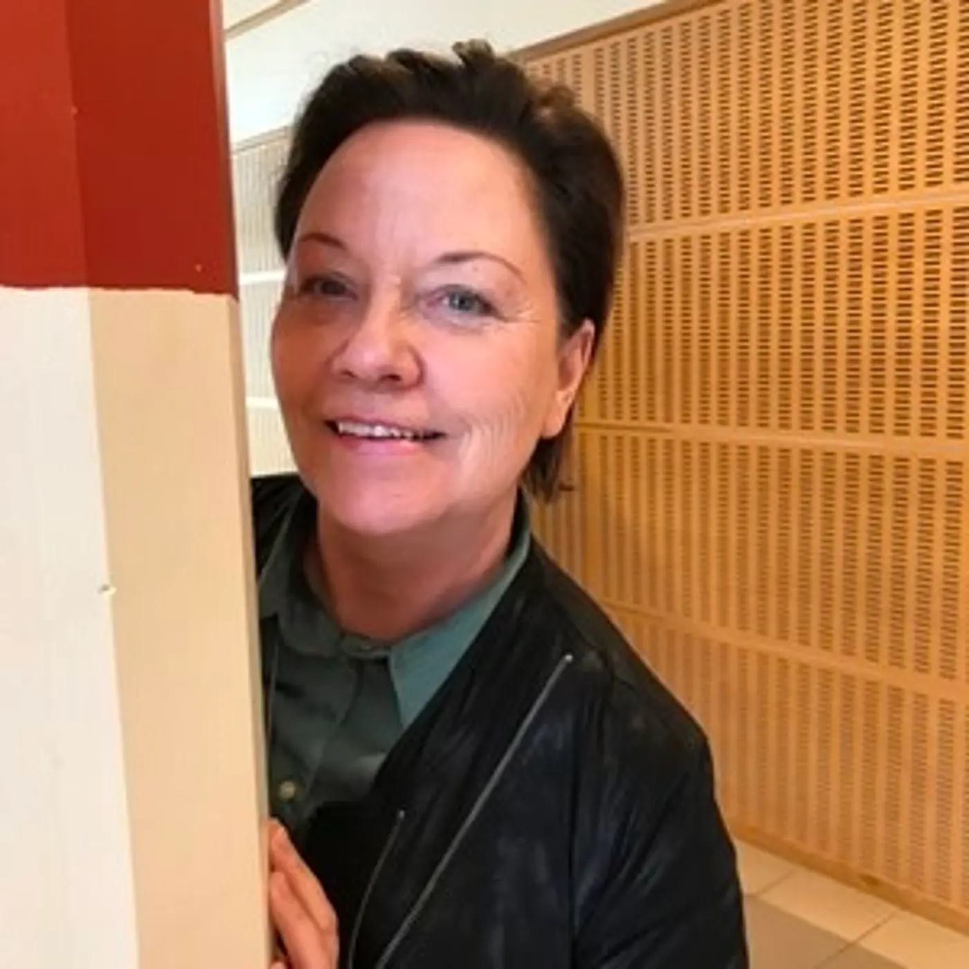 En leende kvinna med kort hår som tittar fram bakom en pelare.