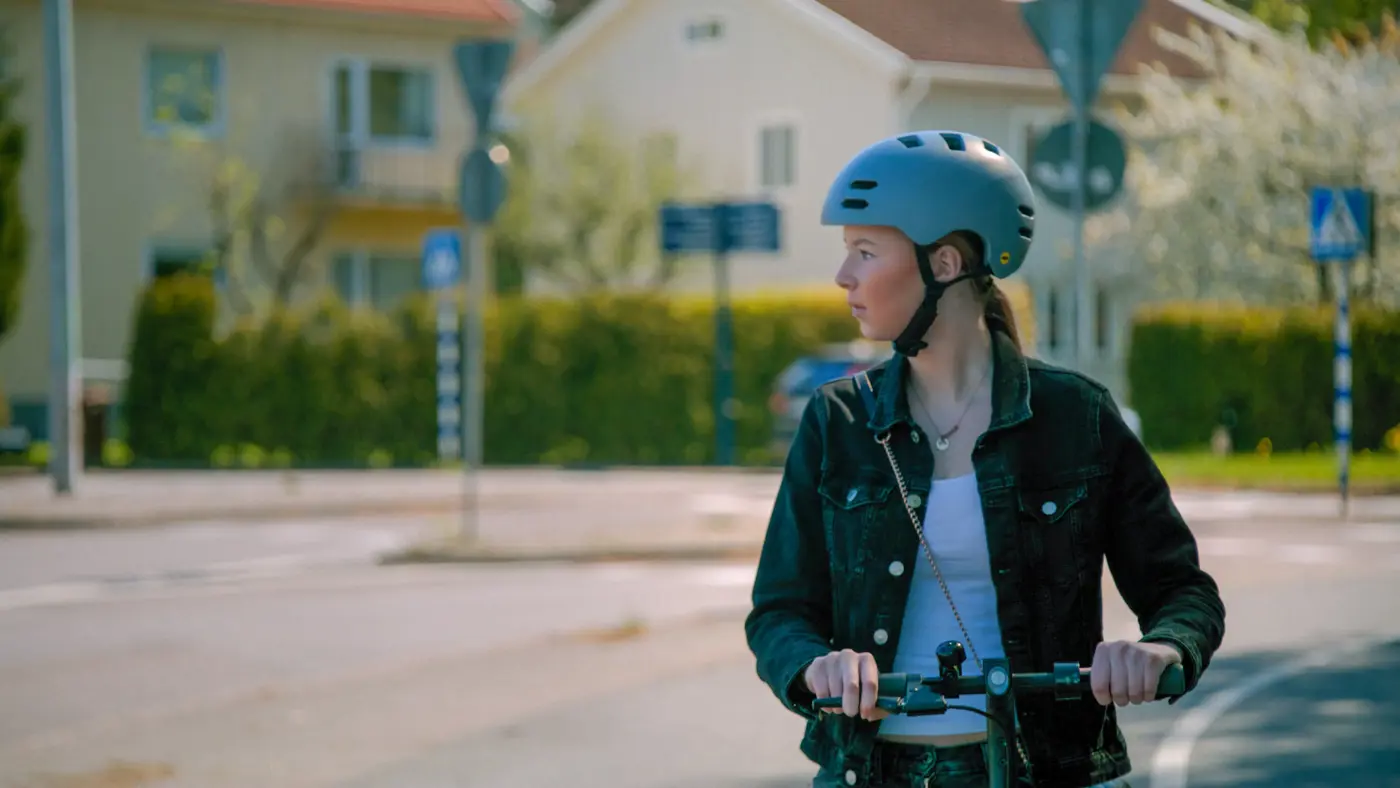En tonåring med hjälp på huvudet kör elsparkcykel på en cykelbana i ett bostadsområde.