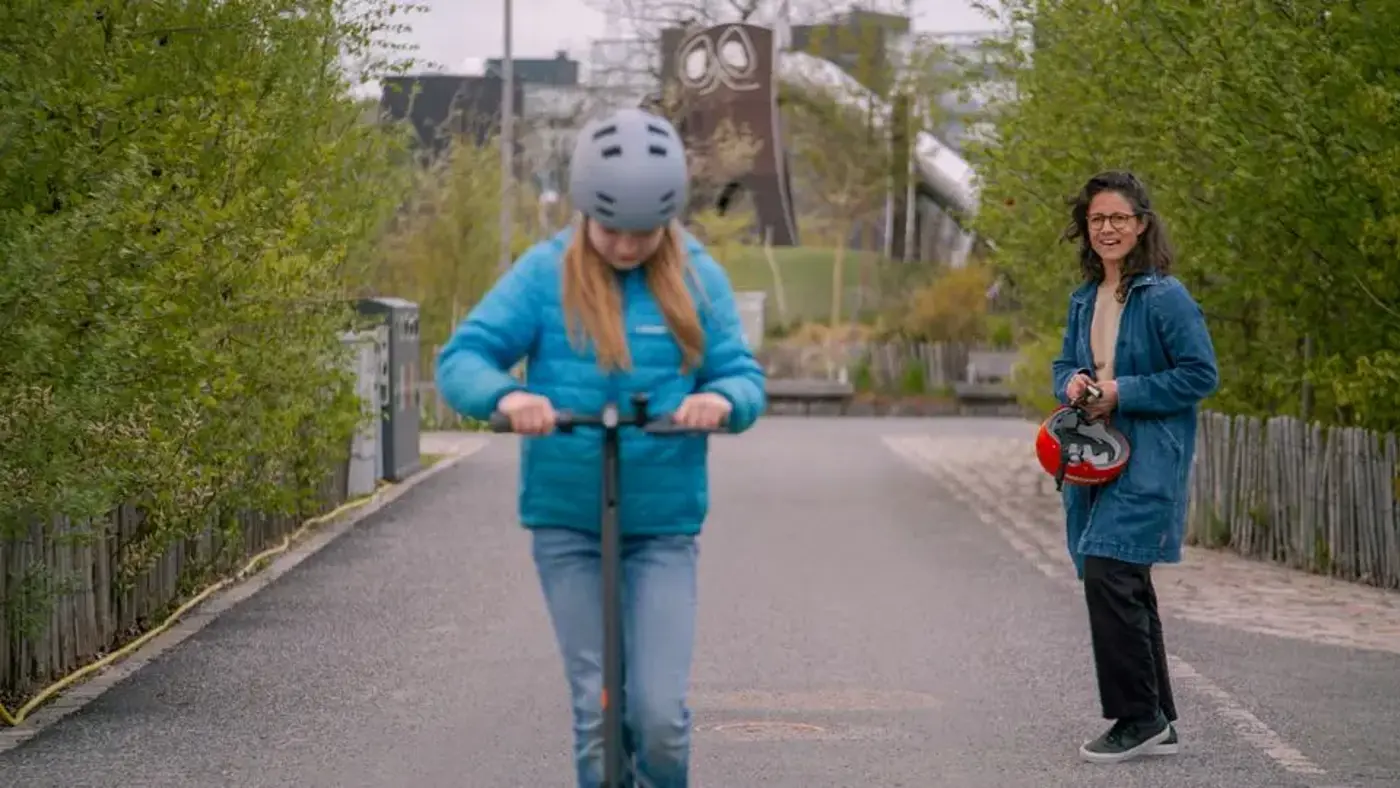 En tonåring med hjälp på huvudet åker elsparkcykel medan en förälder tittar på.
