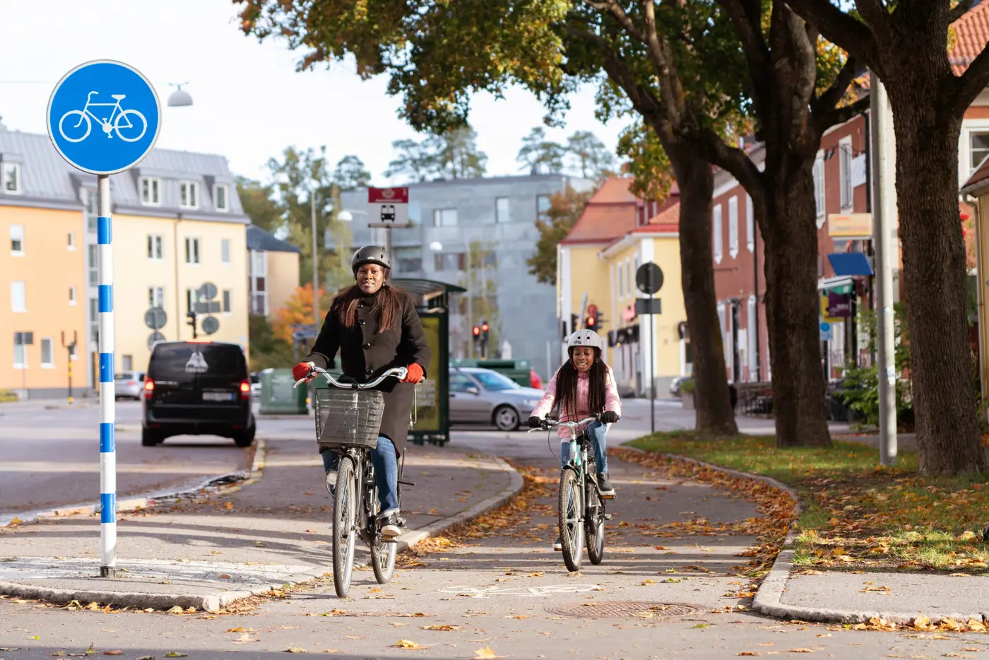 En mamma och ett barn cyklar bredvid varandra på en gång- och cykelbana i stadsmiljö.