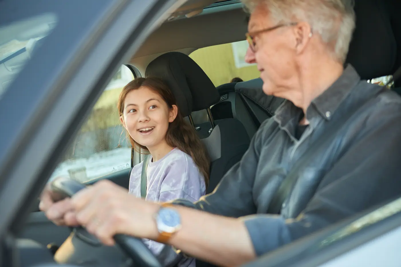 En äldre man kör bil, i passagerarsätet sitter hans barnbarn.