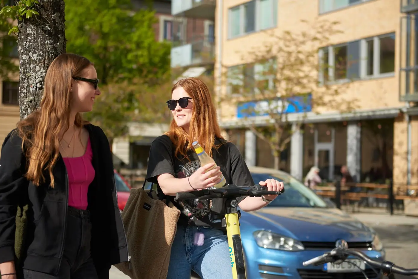 En ung kvinna står på en elsparkcykel med en ciderflaska i handen. Hon pratar med en annan person som står bredvid.