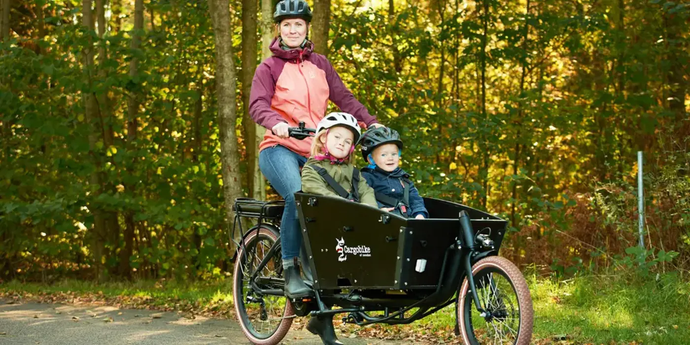 En förälder cyklar på en lådcykel med sina två barn. Alla har hjälm på huvudet.