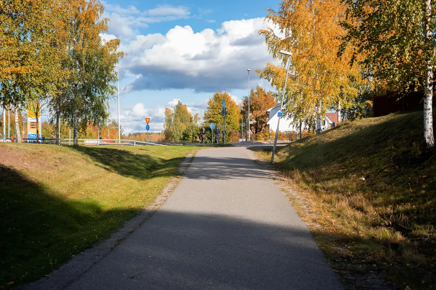 En asfalterad gång- och cykelbana omgiven av gräs och träd varifrån man kan skymta en rondell lite längre fram.