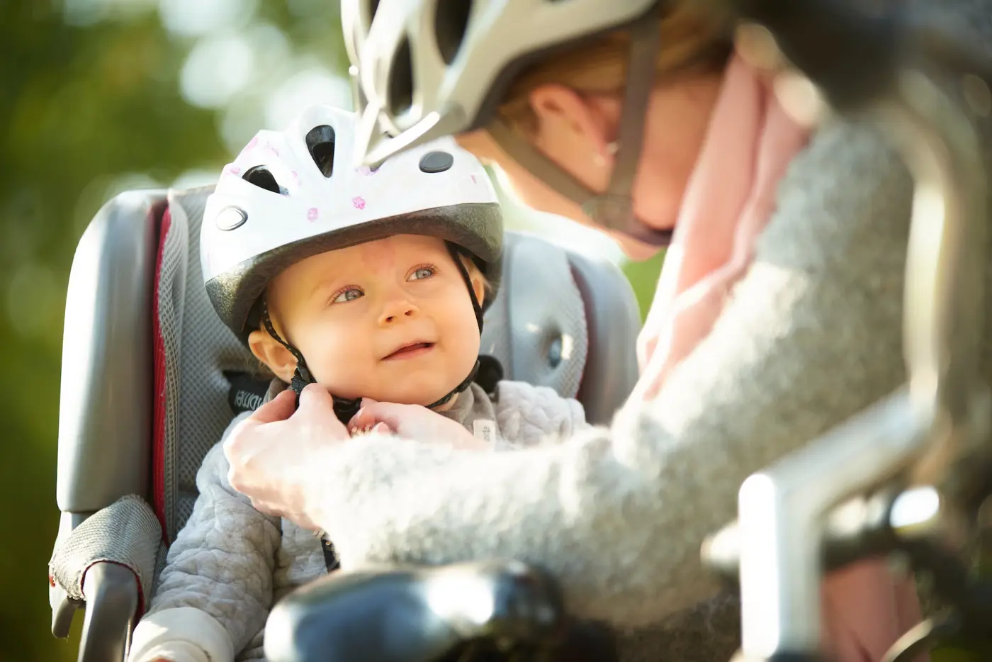 En mamma hjälper sitt småbarn, som sitter i en cykelsits, att spänna cykelhjälmen.