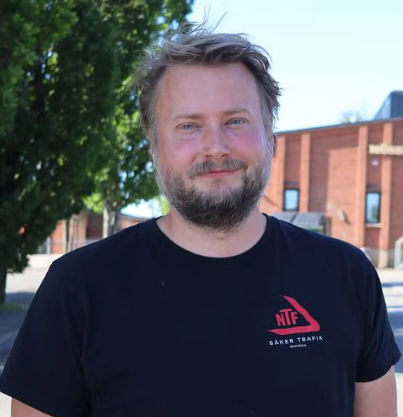 En leende man med svart t-shirt står utomhus vid ett träd.