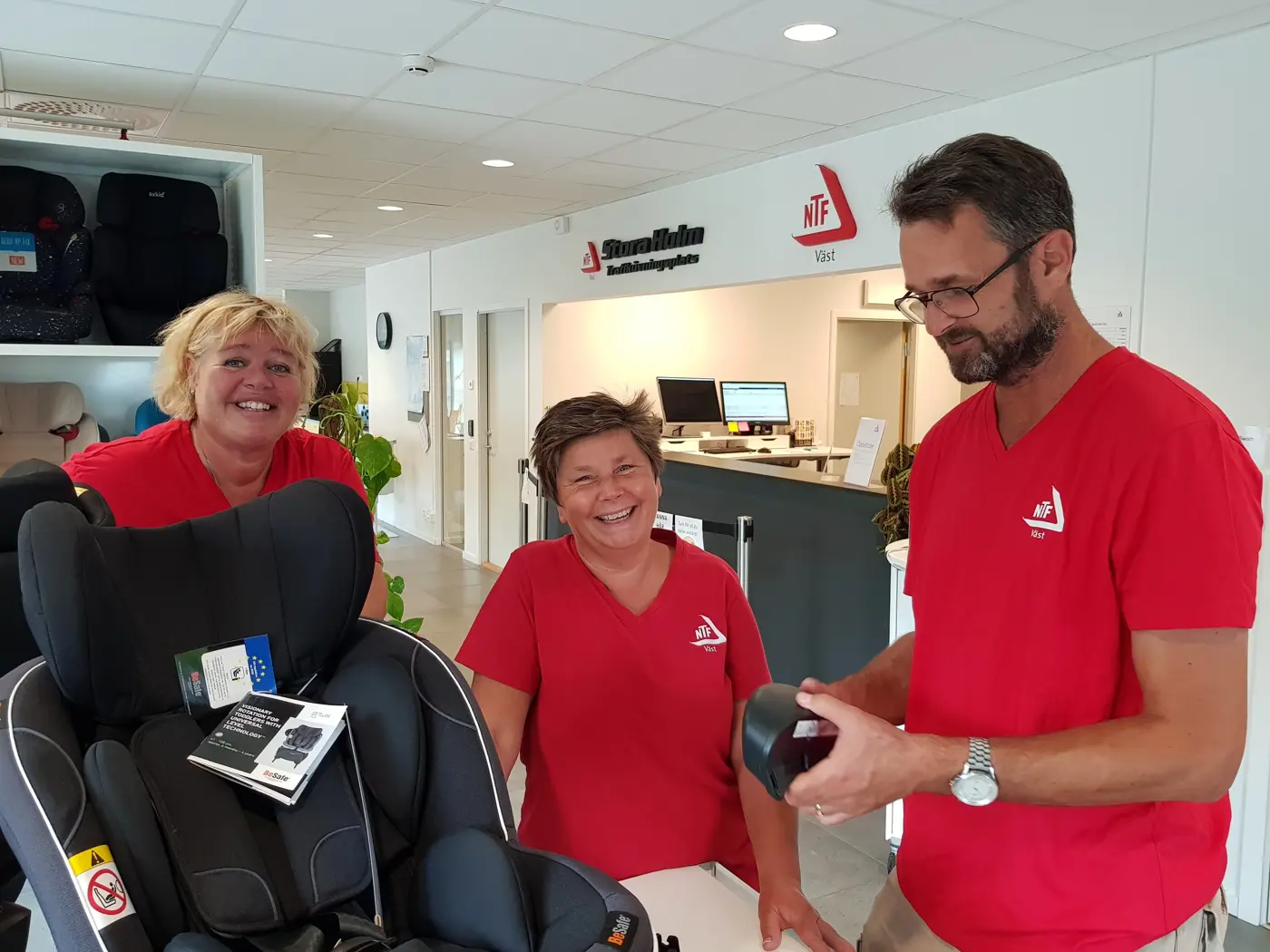 Tre personer står i ett showroom med bilbarnstolar och ler mot kameran.