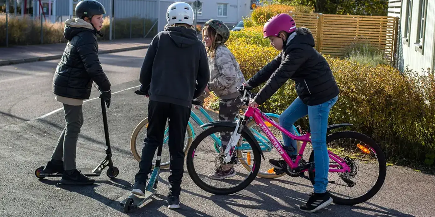 Fyra barn står och pratar på en asfaltsväg. Två sitter på varsin cykel och två står med varsin sparkcykel.