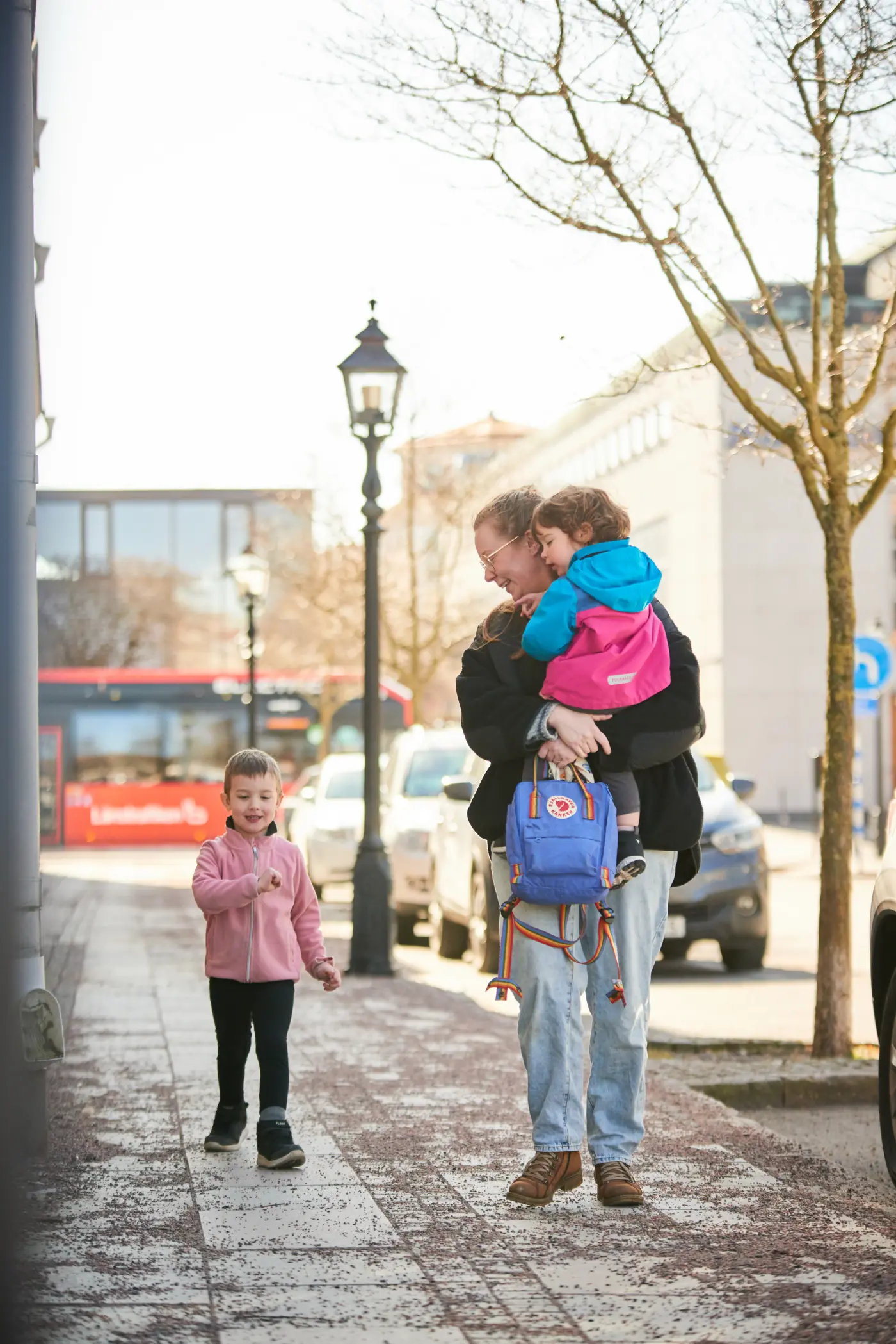 En mamma promenerar med sina två barn. Hon bär ett barn i famnen och det andra barnet går bredvid.