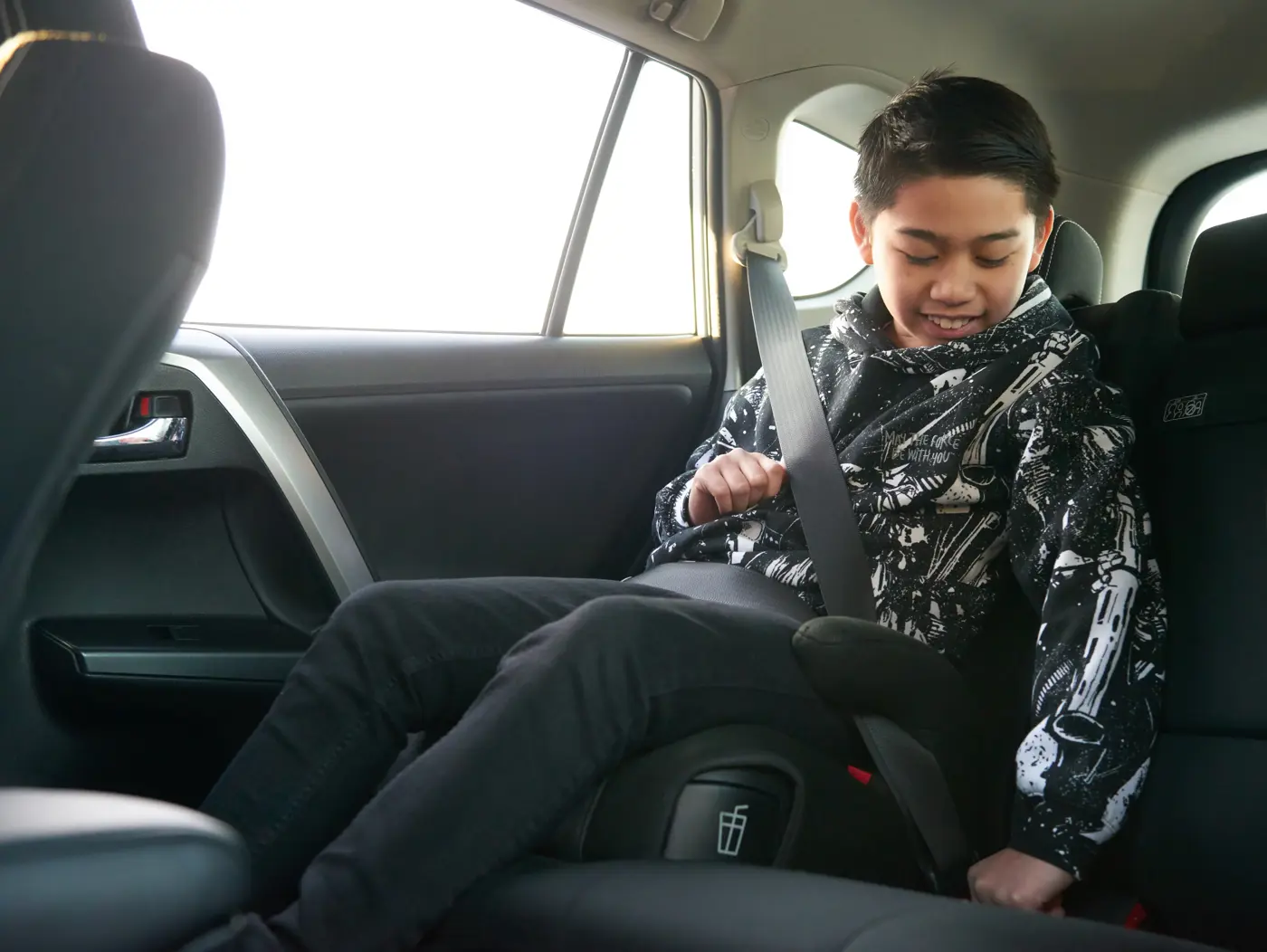 En liten kille som sitter på bälteskudde i bil.