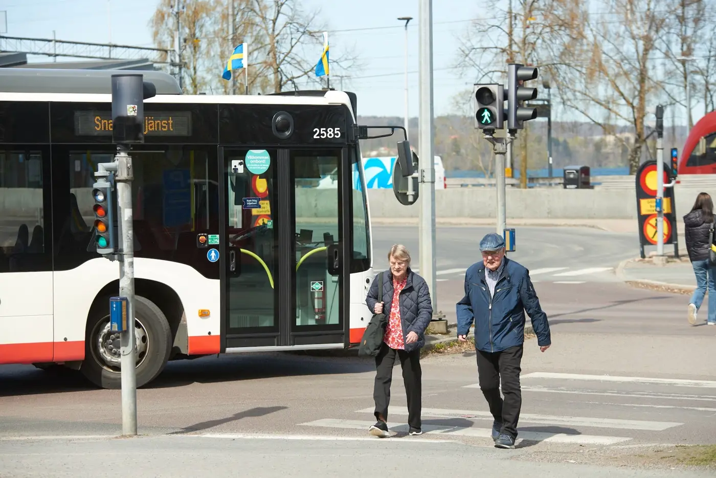 Två äldre personer går över ett bevakat övergångsställe och en buss står och väntar på att få köra.