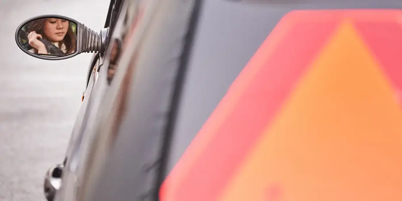 En ung person med blicken nedböjd syns i vänstra sidospegeln på en svart mopedbil.