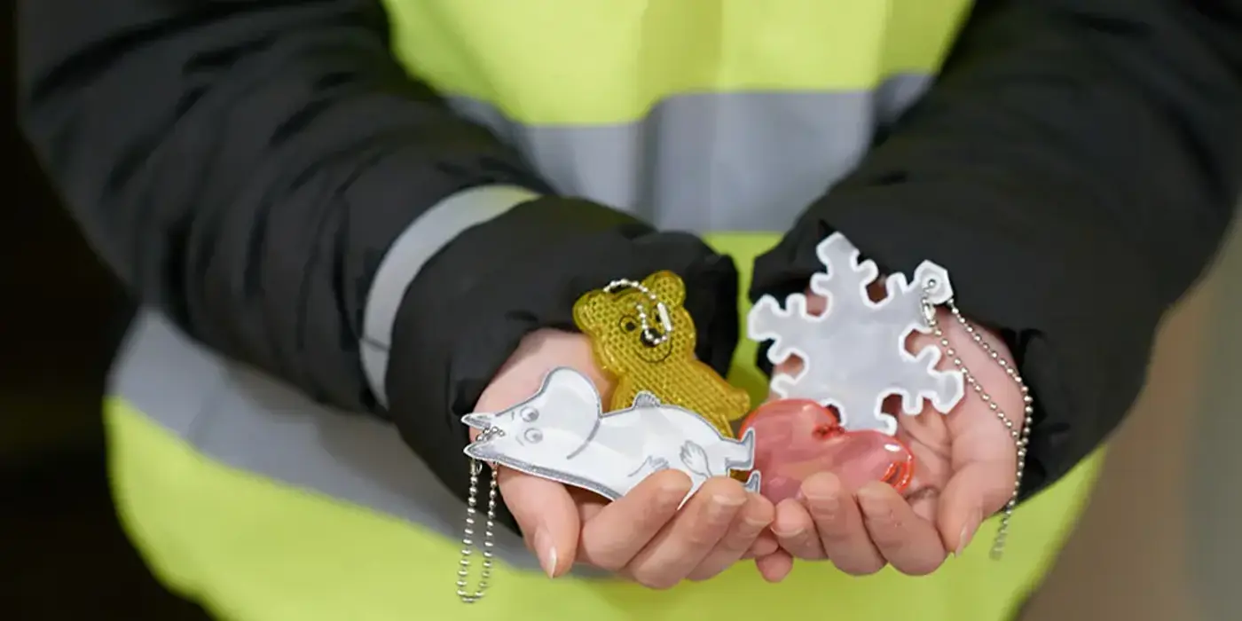 Person med reflexväst och svart jacka håller fram händerna där det ligger fyra reflexer i olika former och färger.