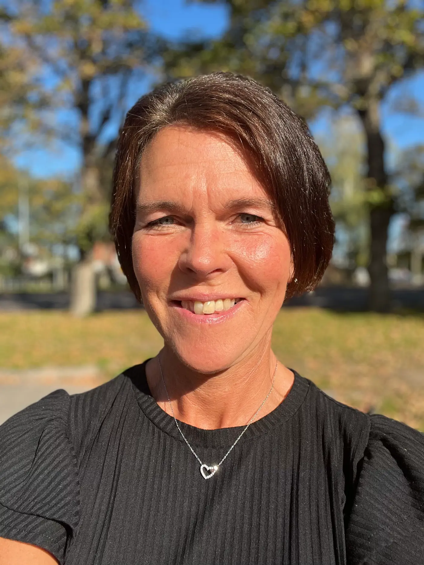 En leende kvinna med kort hår och svart tröja. Hon står utomhus i solljus.