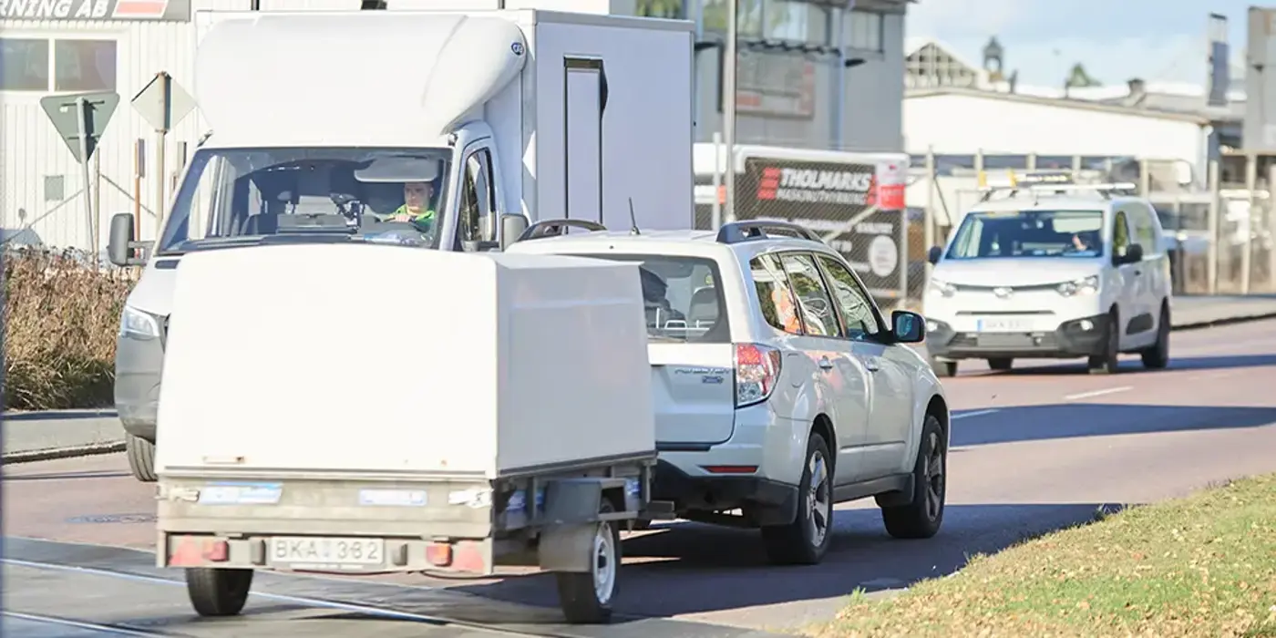 Vit bil kör med släpkärra med kåpa på en väg.