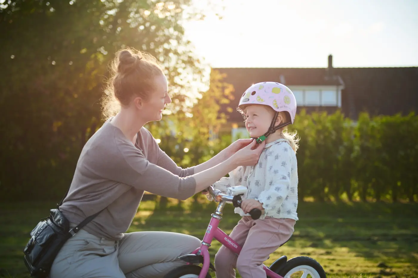 Ett barn sitter på en balanscykel och får hjälp av en förälder att knäppa hjälmen.