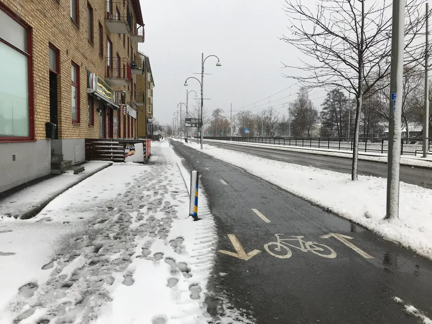 Cykelbana mellan ett lägenhetskomplex och en väg. Vintermiljö med snöslask.