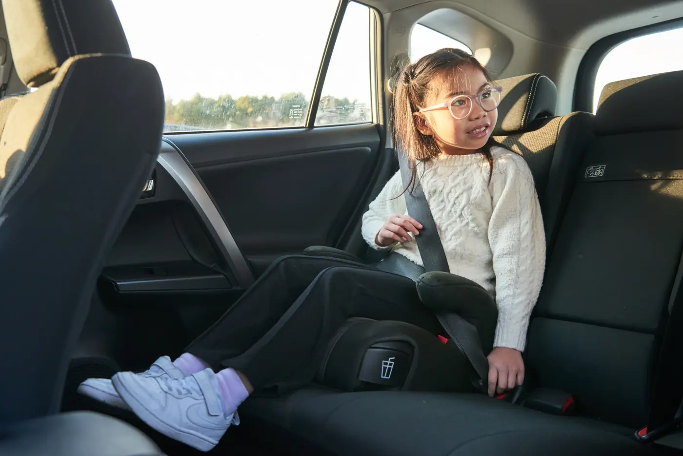 Liten tjej med glasögon som sitter på bälteskudde i bil.