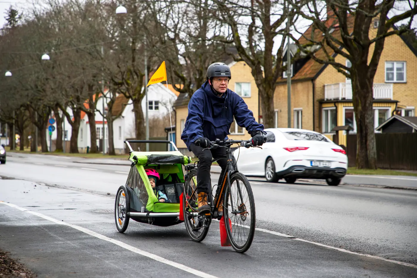En pappa cyklar med en cykelvagn påkopplad, på en cykelbana. Ett småbarn sitter i vagnen.