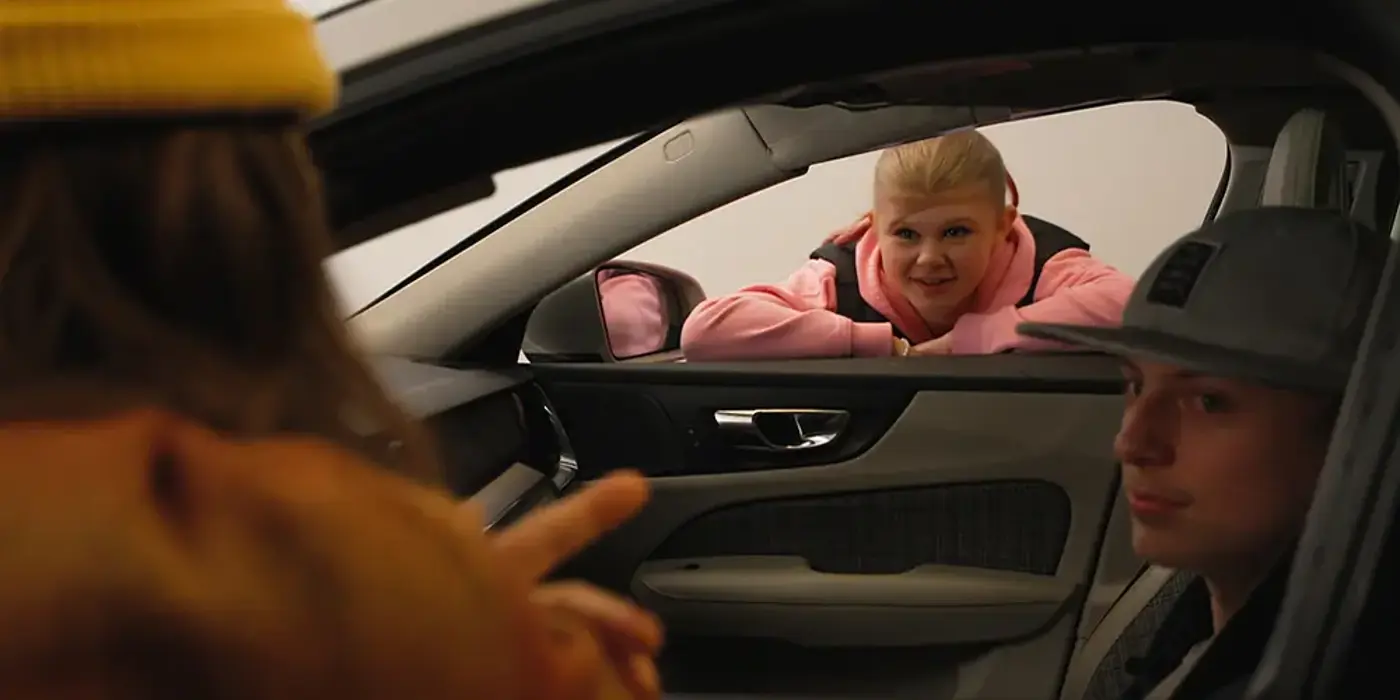 Två personer sitter i en bil och ytterligare en person står lutad mot bilen och pratar med personerna genom passagerarfönstret. Personerna är ungdomar.