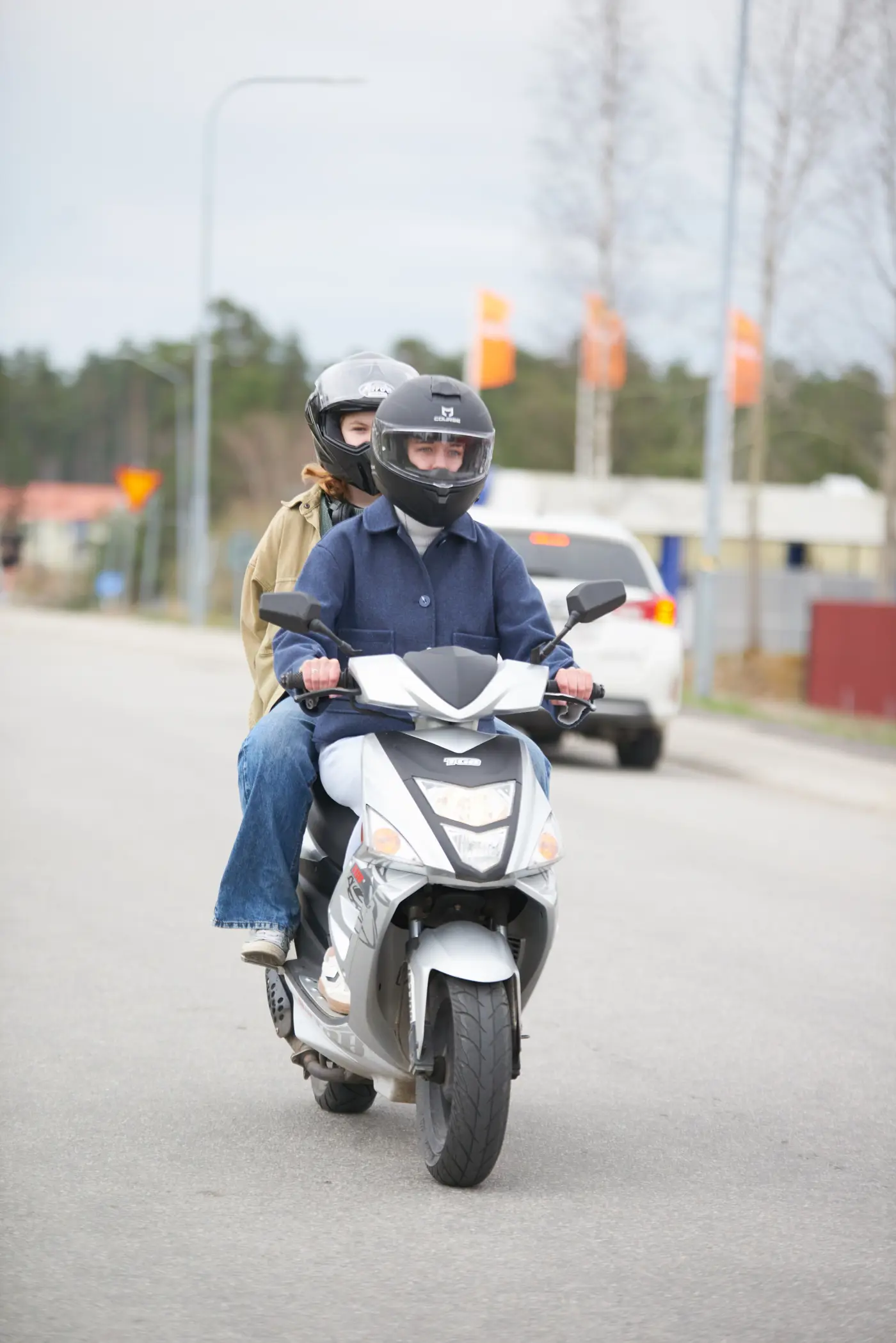 En ungdom skjutsar en annan ungdom på en moped. Båda har hjälm på huvudet.